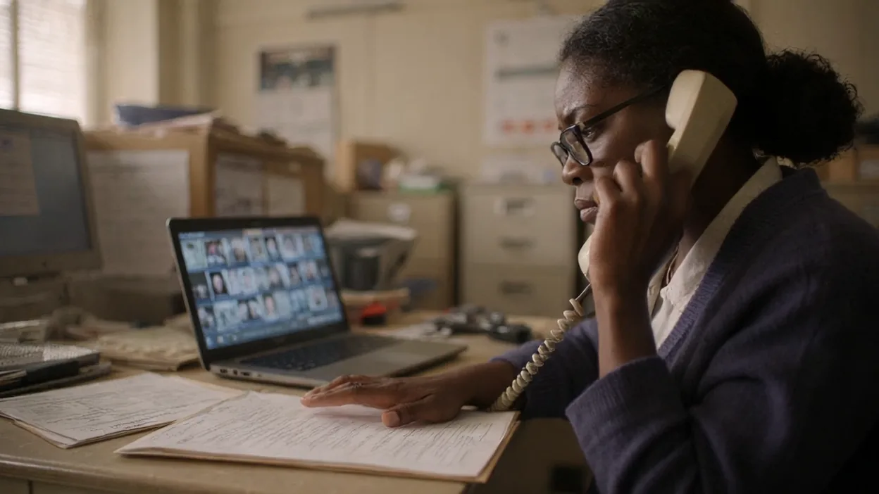 A bookkeeper on a desk phone with one hand on a stack of printed papers, a laptop beside her showing a grid of video-call faces.