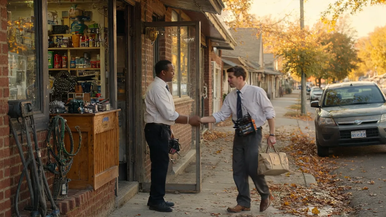 A store owner shakes hands with an arriving technician carrying a tool bag at the door of a brick storefront on an autumn street.