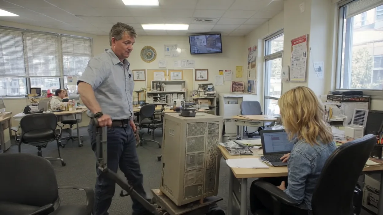 A technician steers a dolly loaded with an old beige tower server through an office while a woman at a nearby desk works on a laptop.