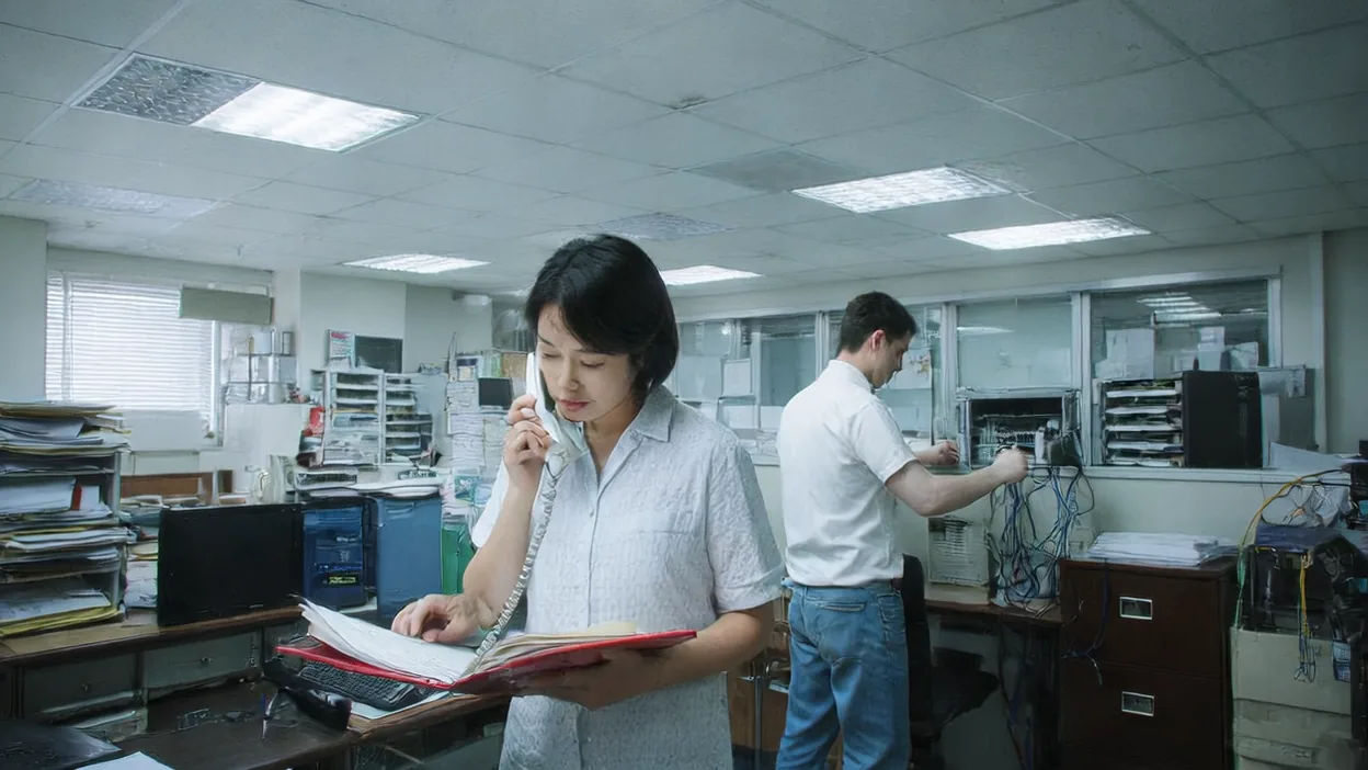 An office manager reads from a red binder while on the phone as a technician unplugs cables from a workstation behind her.