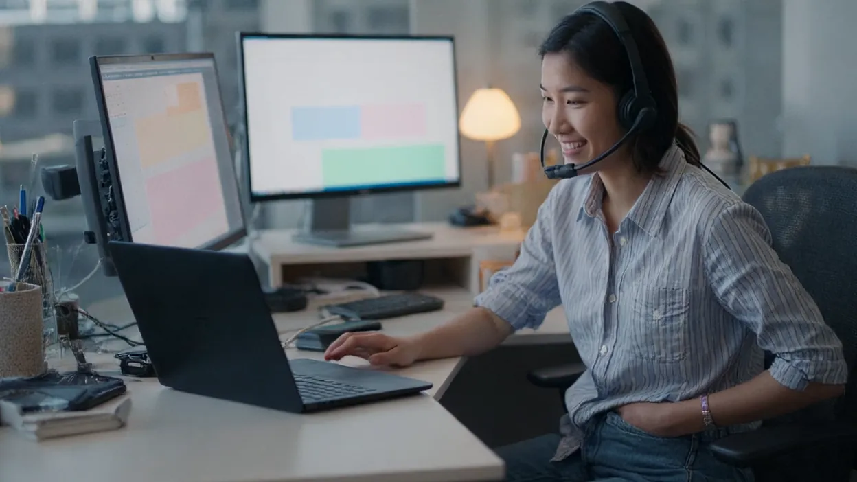 A smiling help desk technician in a headset with a boom mic rests her hand by a laptop, two monitors on the desk behind her.