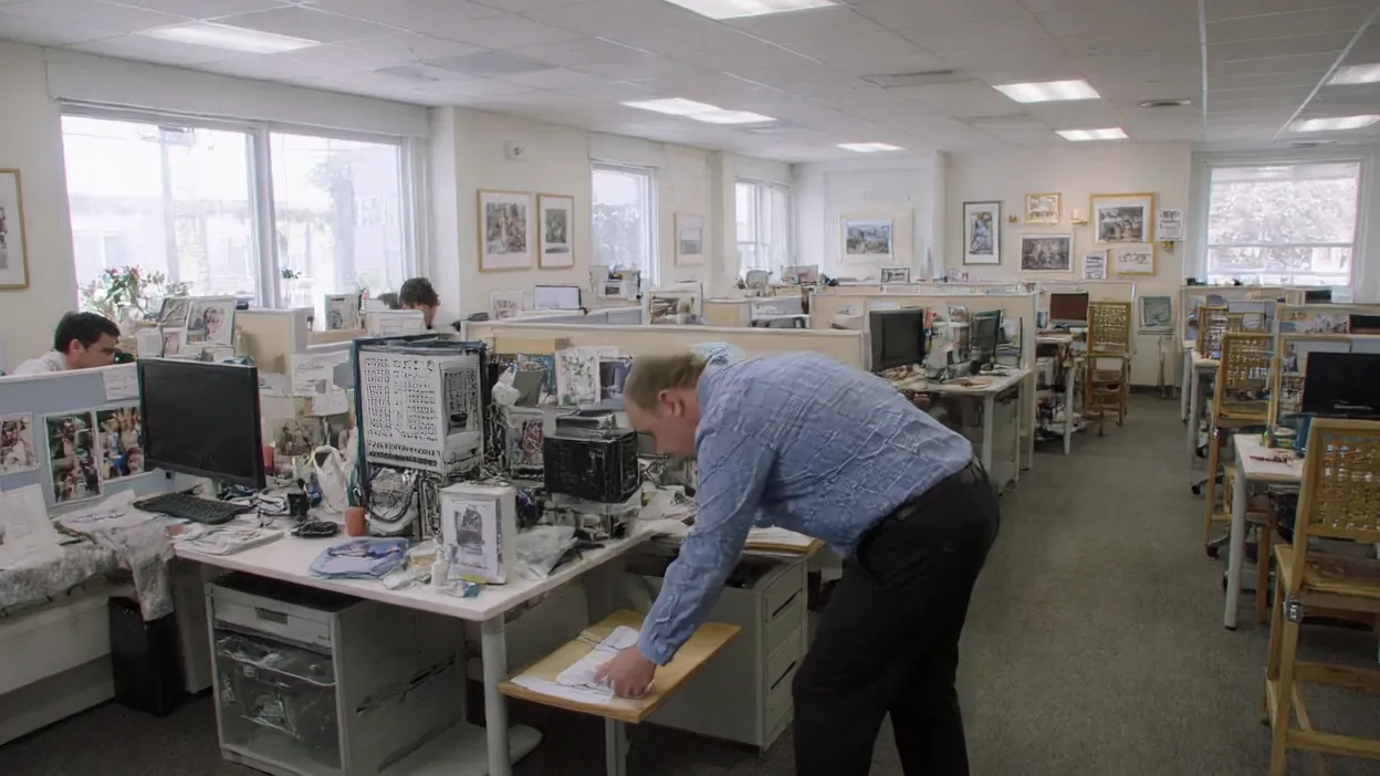 A technician leans over a desk taking notes as he inventories computers in an open-plan office.