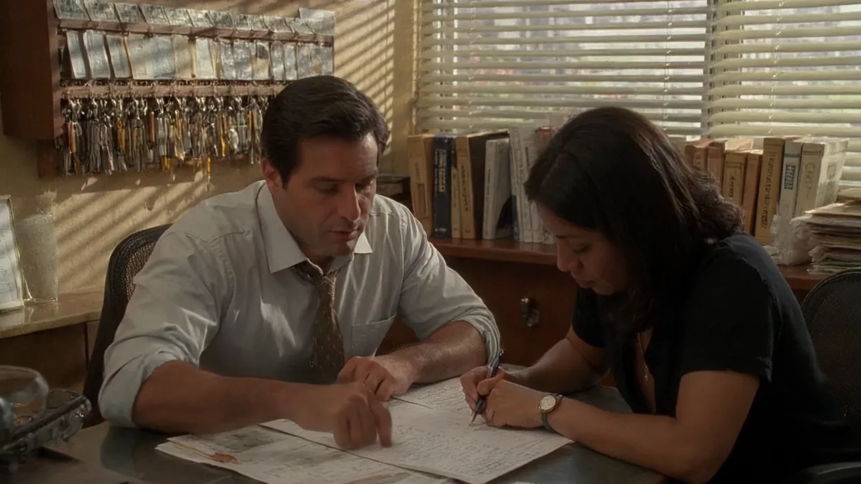 Two people mark up a printed staff list at a desk in a law office, a cabinet of labeled keys on the wall behind them.
