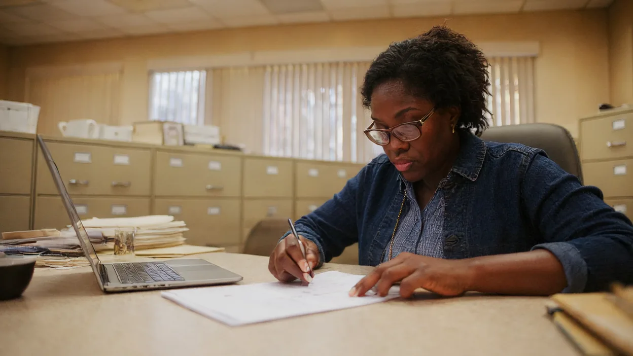 A business owner fills out a paper checklist at a table in an office lined with filing cabinets.