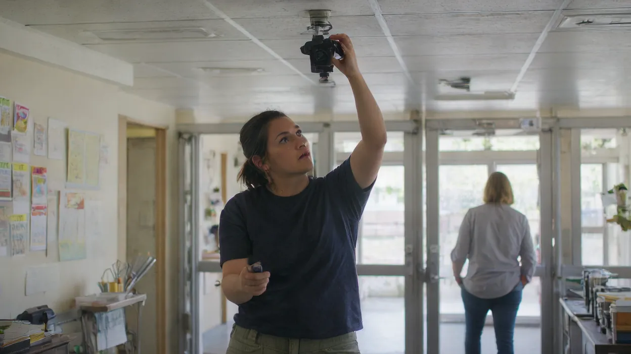 A technician reaches up to adjust a camera mounted on the ceiling of an office entryway while a coworker walks toward the glass front doors.