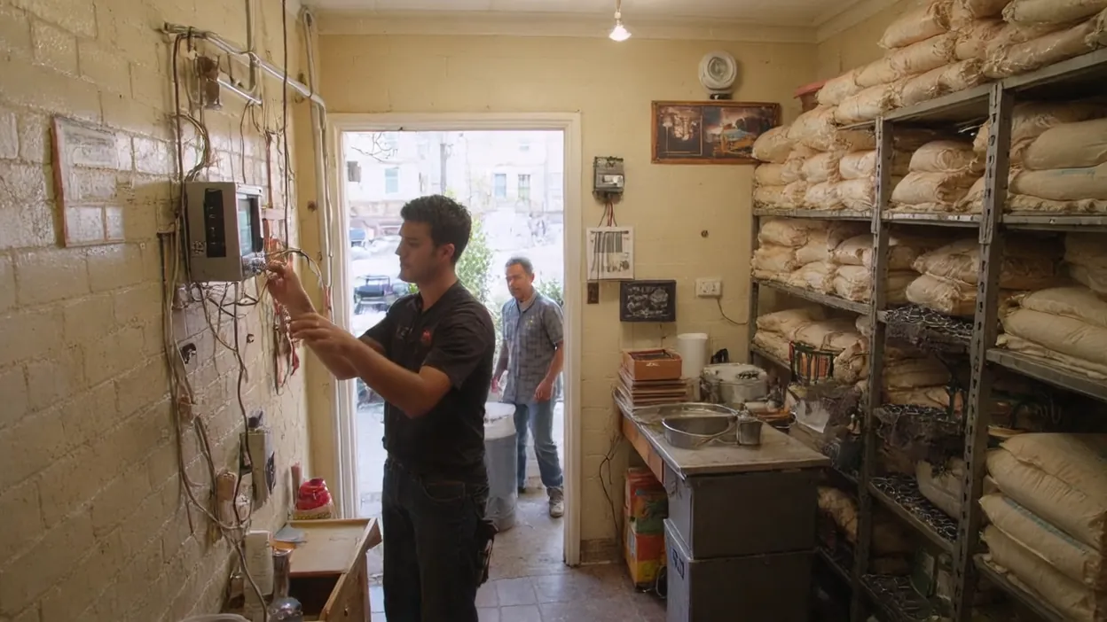 A technician works at a wall of wiring in a bakery stockroom while an installer waits in the doorway.