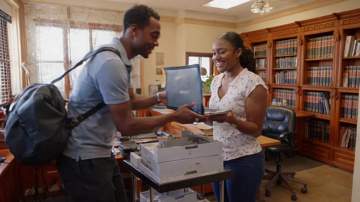 A technician hands a laptop to a new employee in an office lined with wooden bookshelves.