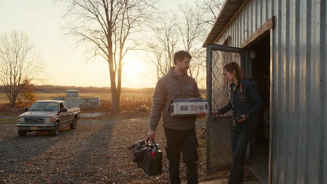 A technician carrying equipment and a tool bag is greeted at the door of a metal farm building at sunset, a pickup truck behind him.
